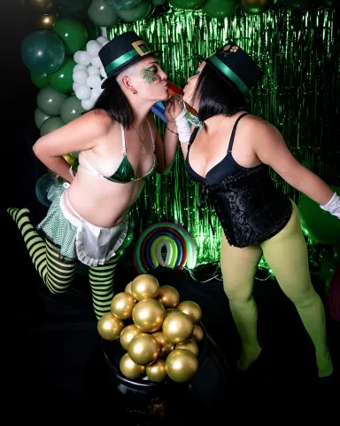 Two women in green St. Patrick's Day outfits and hats kissing in front of a festive green balloon backdrop.