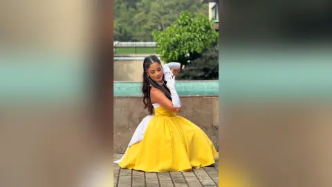 TefyRusso poses outdoors in a large, bright yellow ballgown and white gloves, with her long dark hair styled in a high ponytail.