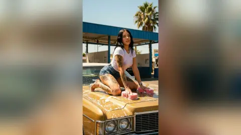 OrianaWill squats on the hood of a gold car, wearing a white t-shirt and denim shorts, showing off her tattoos and legs.