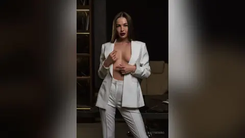 Elegant woman in a white tailored suit posing confidently in a moody, dark luxury interior.