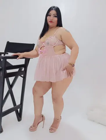 Curvy woman in a pink lace lingerie bodysuit and high heels posing against a white studio background.