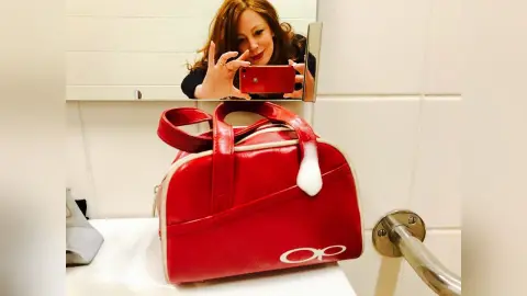 Red leather designer handbag sitting on a white bathroom counter near a woman taking a mirror selfie.