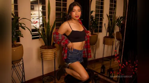 NairaWestbrook poses in a black crop top and denim shorts, wearing an open red plaid shirt while sitting near indoor plants.