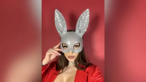 Glamorous woman in a sparkly silver bunny mask and red blazer posing against a red background.