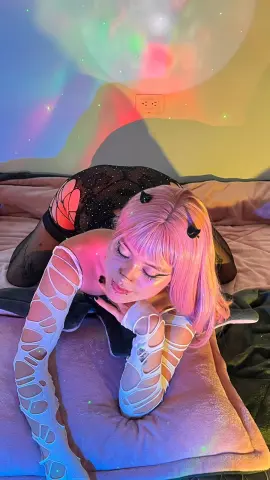 Vakker_Meilee, with pink hair and a black mesh bodysuit, lies on a pink bed under colorful lights.