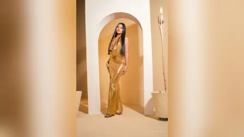 ElieMorgan poses in a long, shiny gold dress standing under a white archway next to a tall torch.