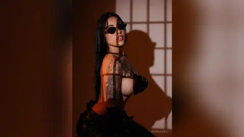 OrianaWill poses in profile wearing sunglasses and a black bra, showing off her long dark hair and arm tattoos against a window shadow.