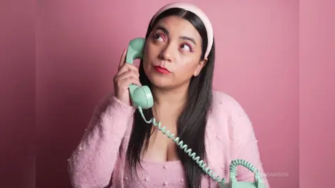 Young woman in pink sweater and headband talking on a vintage mint green telephone against a pink background.