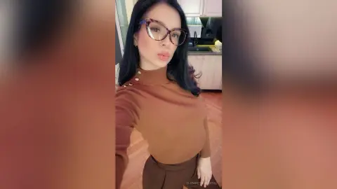 ElizabethChantal — Young woman in a brown turtleneck sweater and glasses posing in a modern kitchen with a confident mood.