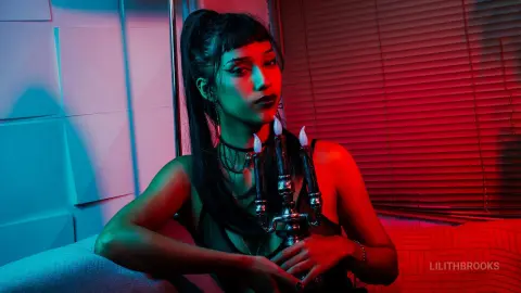LilithBrooks poses in a dark room with red and blue lighting, wearing a black lace top and a black choker.