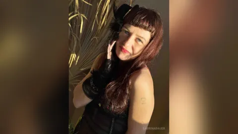 SabrinaBauer — SabrinaBauer smiles in a black lace outfit and hat while posing near palm leaves.