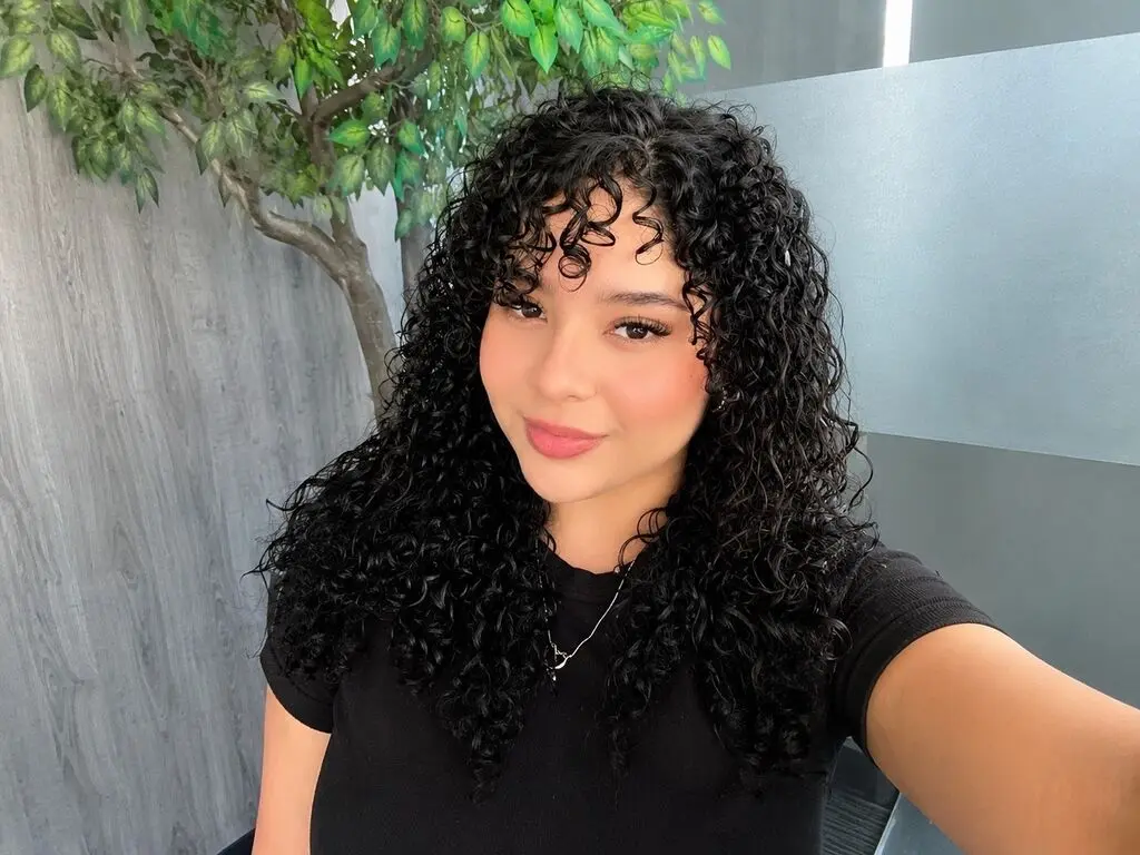 EmilyJune poses with her long, curly black hair and a black t-shirt against a background of green leaves and a grey wall.