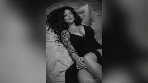 Glamorous woman with curly hair in a black dress lying on a bed in a moody black and white portrait.