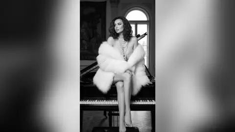 Elegant woman in a white fur coat and pearls sitting on a grand piano in a luxurious vintage room.