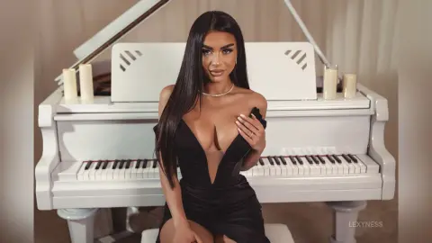 Elegant woman in a black plunging neckline dress sitting by a white grand piano in a bright room.