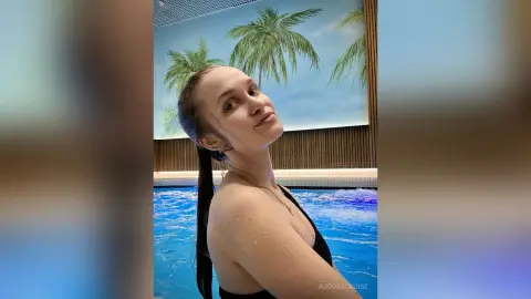 AuroraCruise poses in a black bikini or top by a blue swimming pool, showing her wet skin and long dark hair tied back.