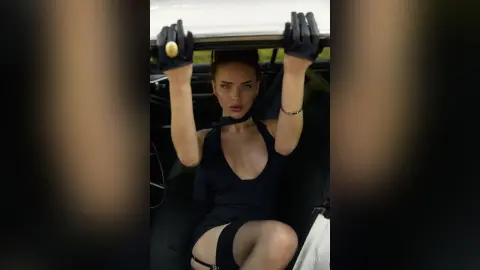 AnjeNoir wears a low-cut black dress and black gloves while sitting in a car.