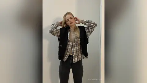 Blonde woman in a plaid shirt and black vest posing confidently in a minimalist indoor setting.