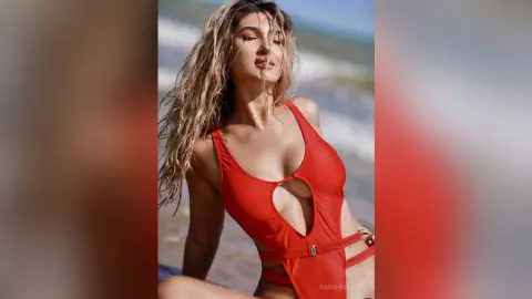 Blonde woman in a red cutout swimsuit posing on a sunny beach with a relaxed summer mood.