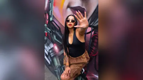 ChloeRiveiro — Stylish woman in a black bodysuit and sunglasses posing confidently in front of vibrant urban street art.