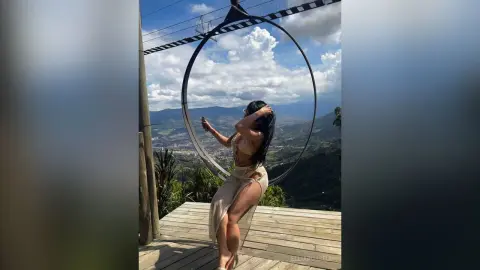 Woman in a beige cutout outfit posing on a giant swing overlooking a scenic mountain valley under a blue sky.