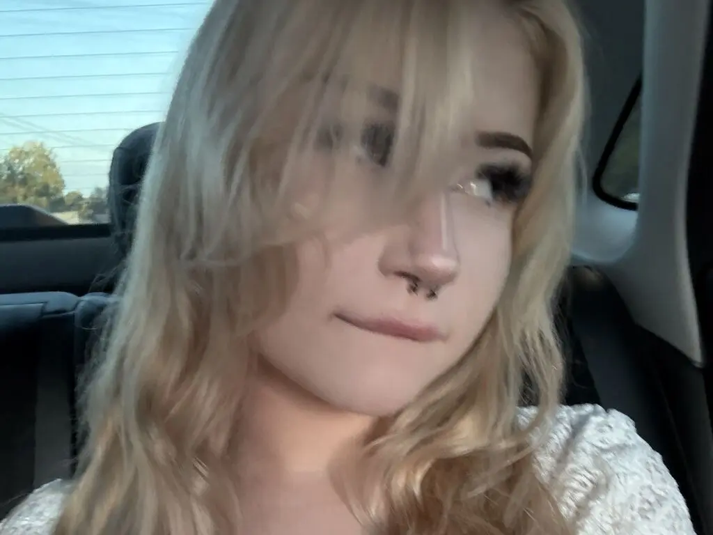 Blonde woman with nose ring wearing a white knit sweater sitting in a car on a sunny day.