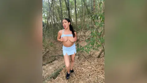 Young woman in a light blue summer outfit posing playfully in a lush, sunlit forest setting.