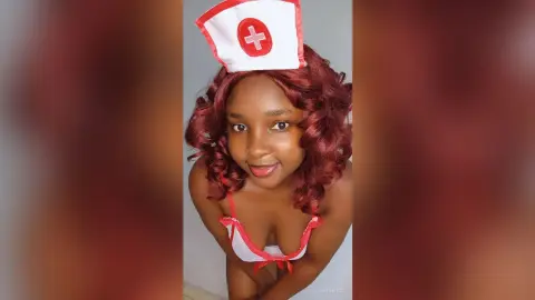 Smiling Black woman in a red and white nurse costume posing against a plain wall.