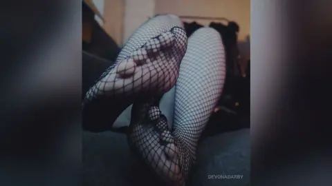 Close-up of feet wearing black fishnet tights resting on a dark sofa in a moody, dimly lit room.