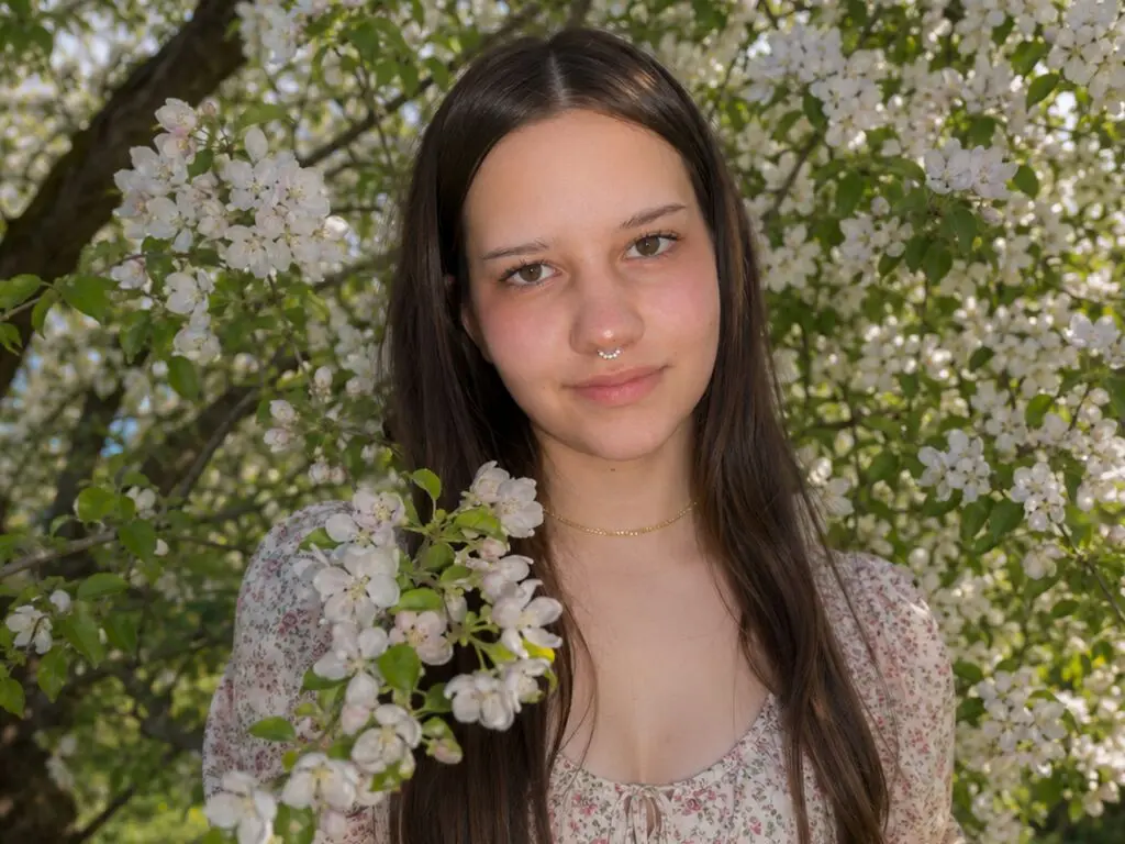 Young woman in a floral dress smiling amidst blooming white apple blossoms in a sunny spring garden.