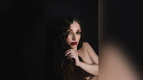 Elegant woman with wavy dark hair and red lipstick posing against a dark moody background.