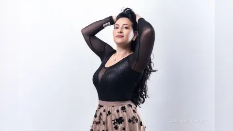 Elegant woman in a black sheer bodysuit and floral skirt posing confidently against a white studio background.
