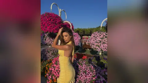 Young woman in a yellow summer dress posing amidst vibrant pink hanging flower baskets in a sunny garden.