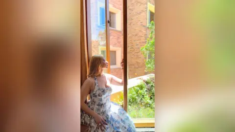 Blonde woman in a floral summer dress looking out a window of a modern brick apartment building.