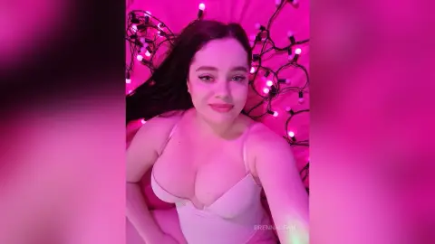 Young woman in white lingerie lying on pink bedding surrounded by glowing pink fairy lights.