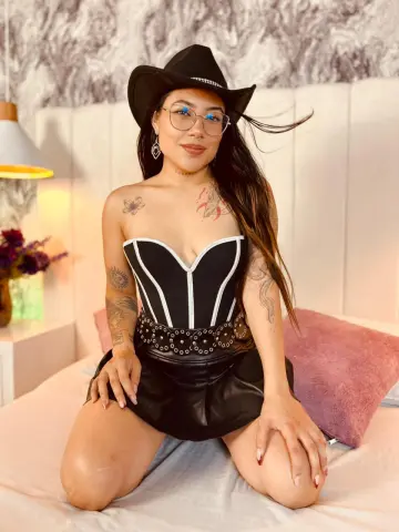 Young woman in a black corset and mini skirt sitting on a bed in a bright, modern bedroom.