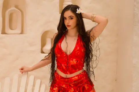Young woman in a red floral summer outfit posing against a white Mediterranean-style wall in bright sunlight.