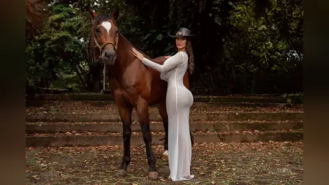 Elegant woman in a white maxi dress posing with a brown horse in a lush, leafy forest setting.