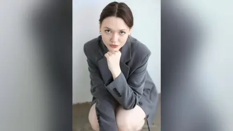 Elegant woman in a grey oversized blazer posing with a thoughtful expression against a minimalist white background.