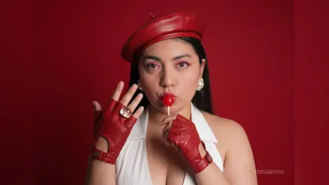 Stylish woman in a red beret and white top eating a red lollipop against a bold red background.