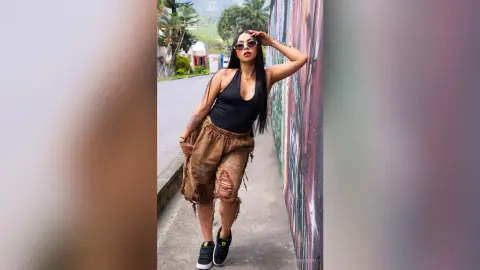 ChloeRiveiro — Stylish woman in a black bodysuit and distressed brown shorts posing against a colorful wall in bright sunlight.