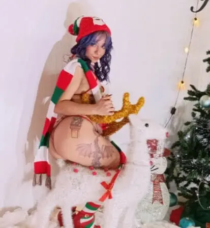Woman in festive red hat and scarf sitting on a white reindeer decoration near a Christmas tree.