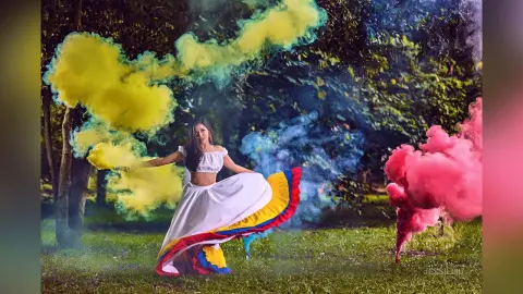 JessieLiu stands in a grassy forest setting wearing a white off-the-shoulder crop top and a long white skirt with a colorful red, yellow, and blue trim, surrounded by thick clouds of yellow, blue, and pink smoke.