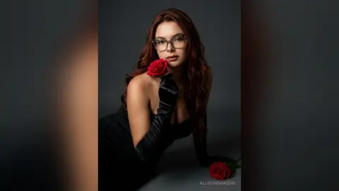 Elegant woman in a black satin dress and gloves holding a red rose against a dark studio background.