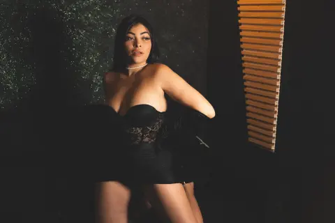 Elegant woman in a black lace corset and pearl necklace posing in moody, low-light indoor setting.