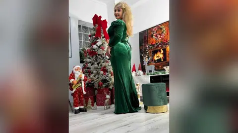Blonde woman in elegant green velvet gown standing by a decorated Christmas tree in a cozy festive living room.