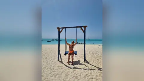 Woman in orange bikini relaxing on a wooden swing at a sunny tropical beach with turquoise ocean water.