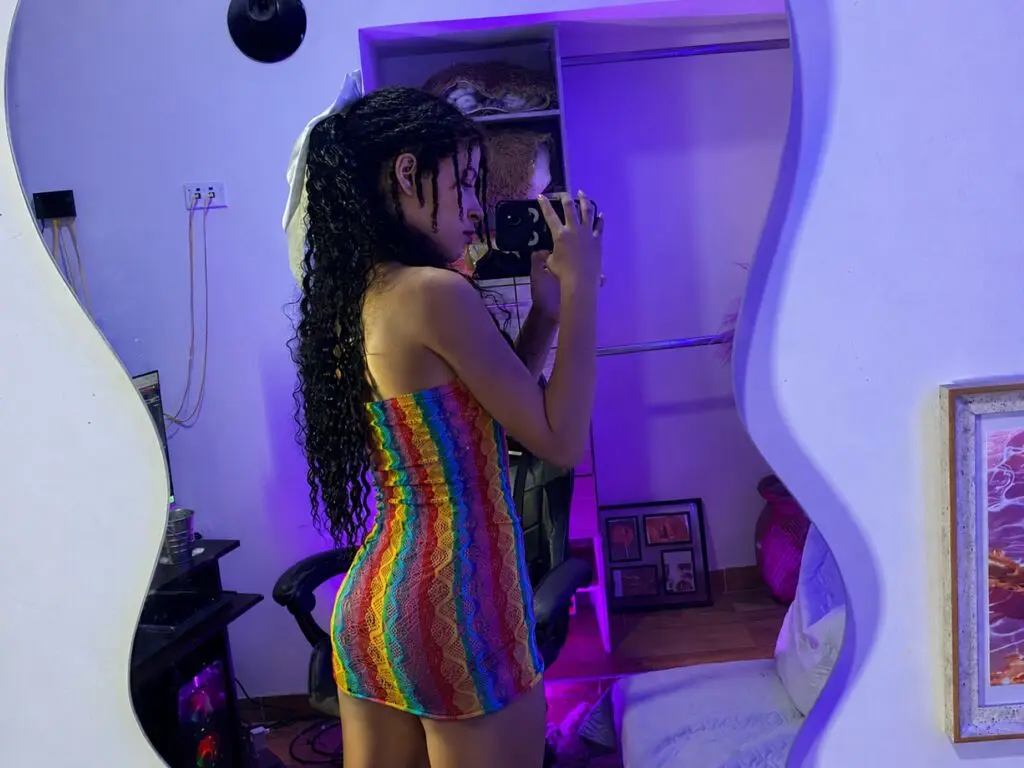Young woman in a colorful rainbow crochet mini dress taking a mirror selfie in a purple lit bedroom.