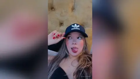 Young woman in a black Adidas cap and black top making a playful expression in a cozy indoor setting.