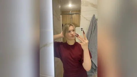 Young woman in a maroon t-shirt taking a casual mirror selfie in a modern bathroom.
